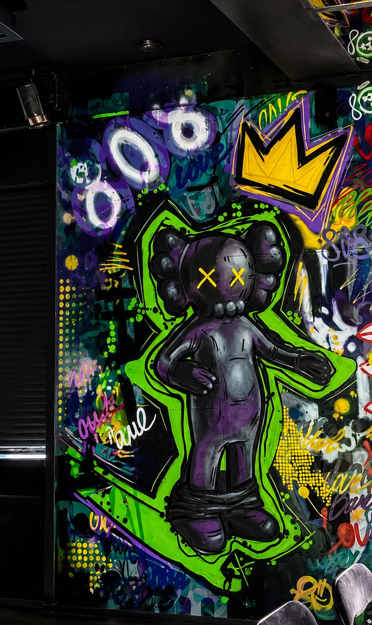 Neon green and violet vinyl-toy character mural with crown and graffiti tags, storefront at night